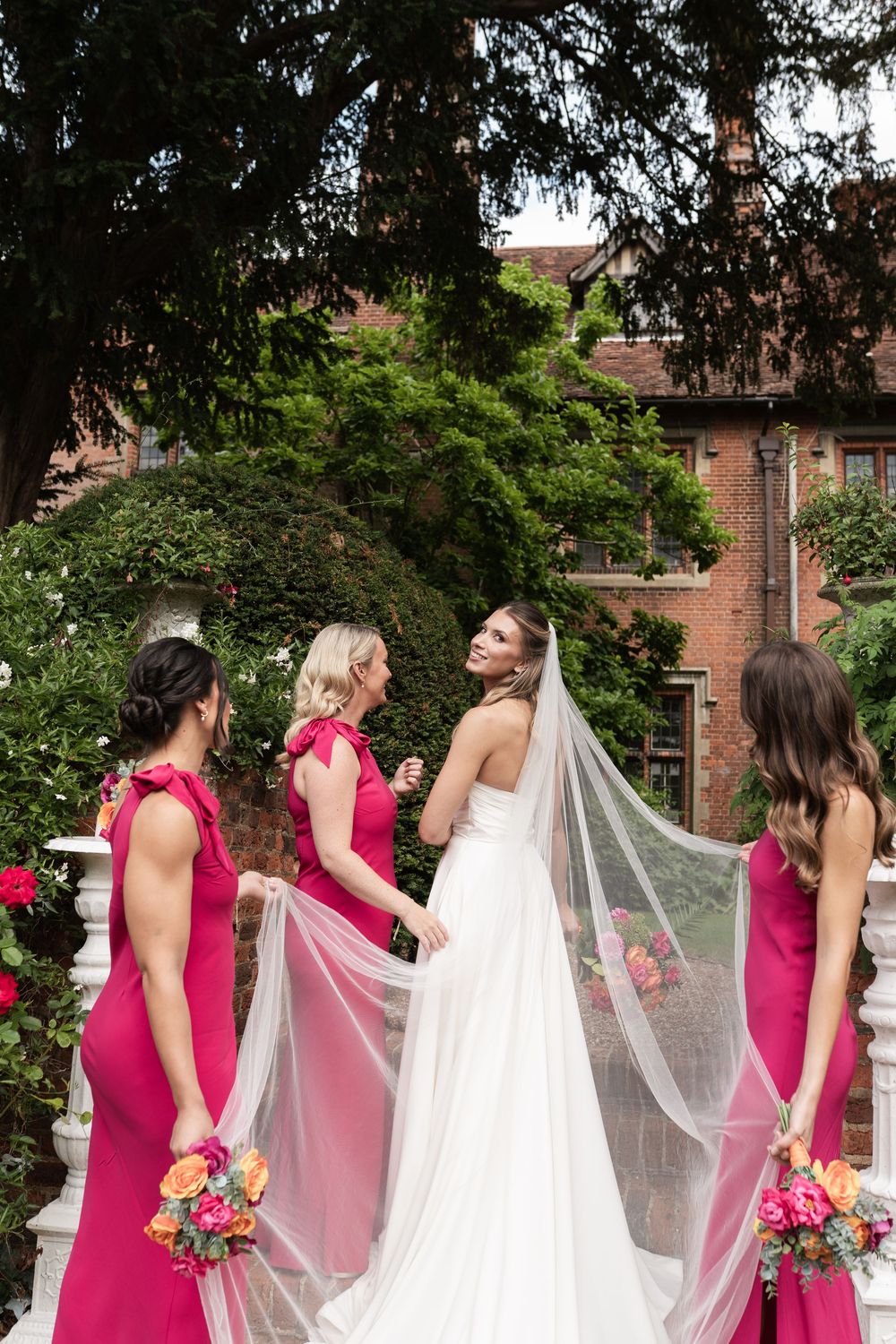 A row of bridesmaids in pink dresses help adjust a long white wedding veil in a garden setting.