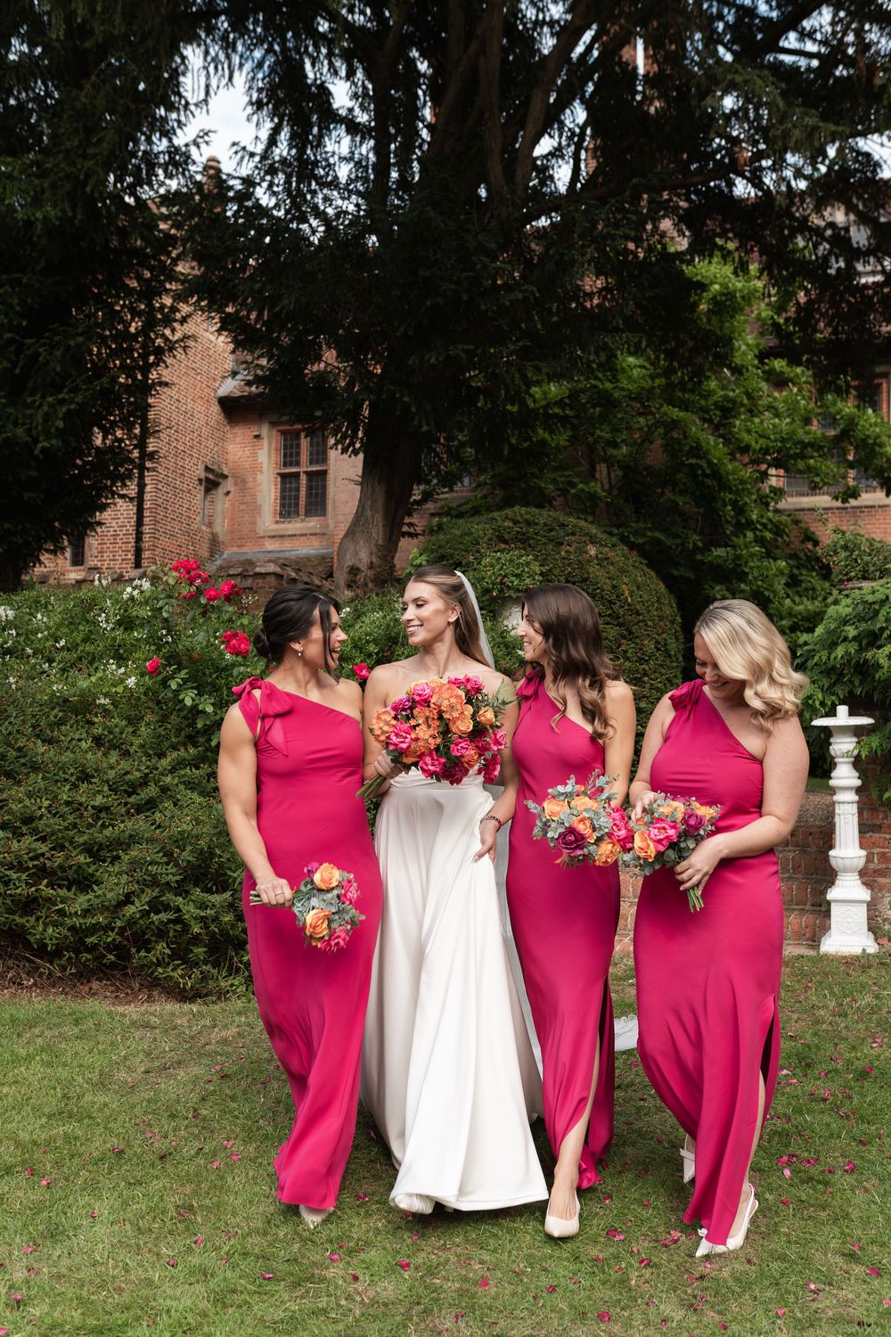 A wedding party in bright pink dresses and white gown poses playfully in a garden setting near a brick building.
