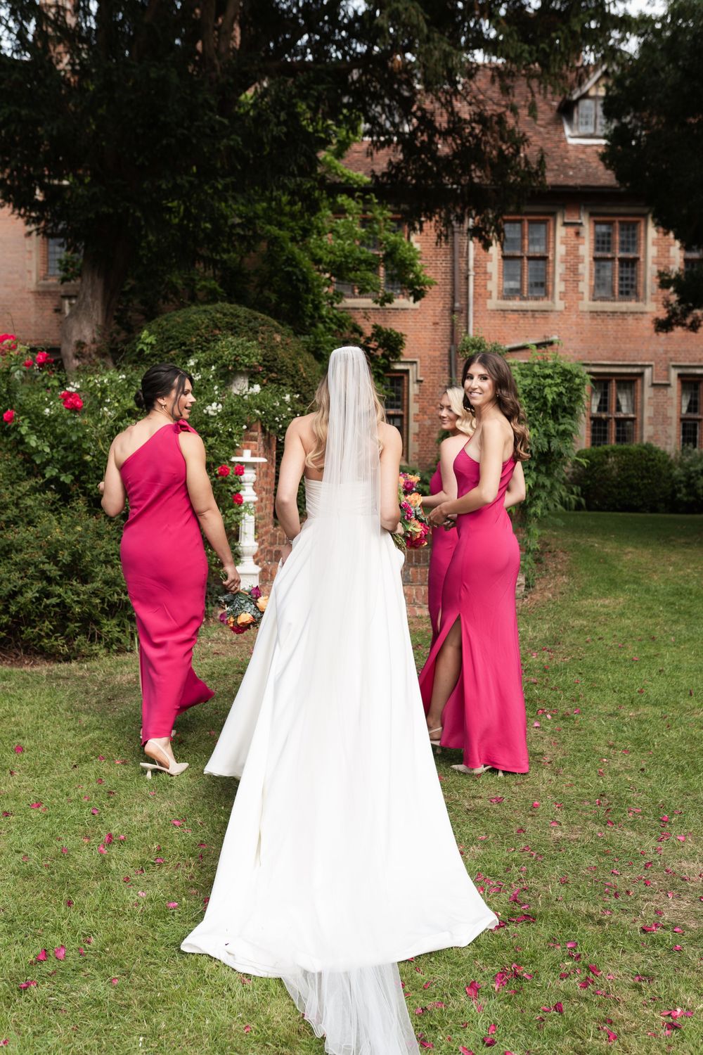 A wedding party stands in a garden with a historic brick building and pink flowers in the background.