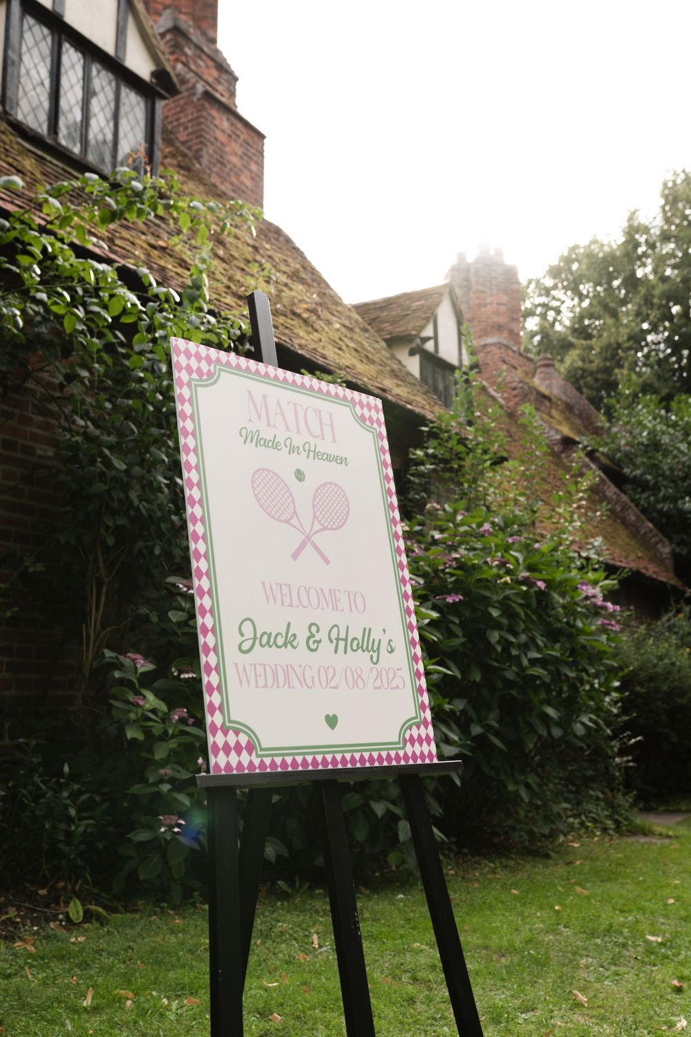 A rustic wooden wedding sign with pink gingham details stands on a black easel in front of ivy-covered brick walls.