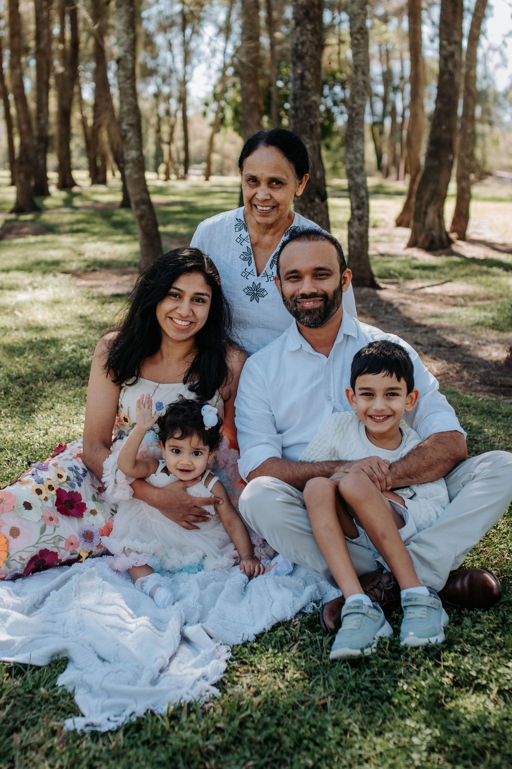 A family group sits together on blankets in a sunlit wooded area on a warm day outdoors.