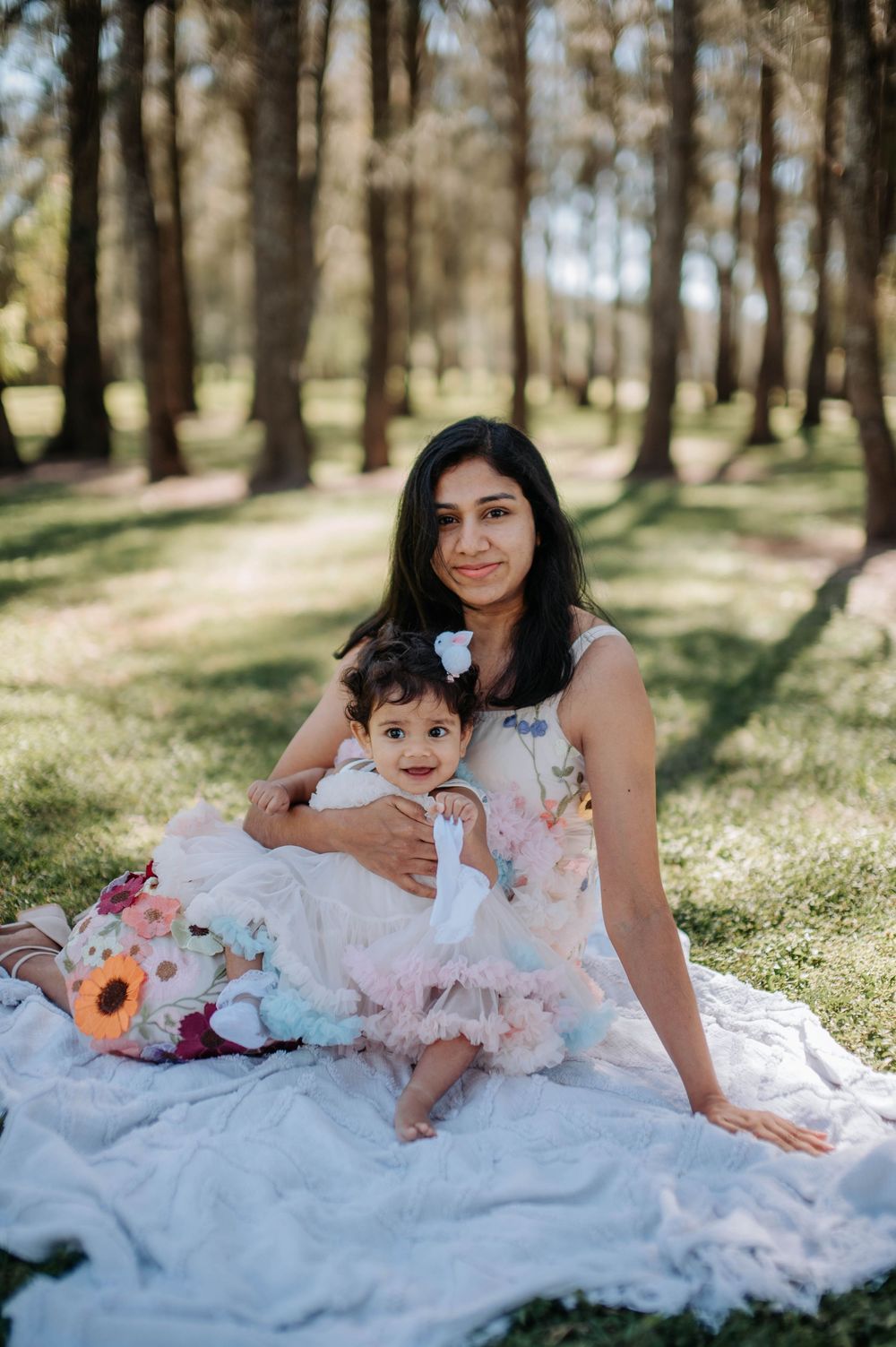 A mother and child sit together on a white blanket in a sunny wooded area during a spring outdoor photo session.