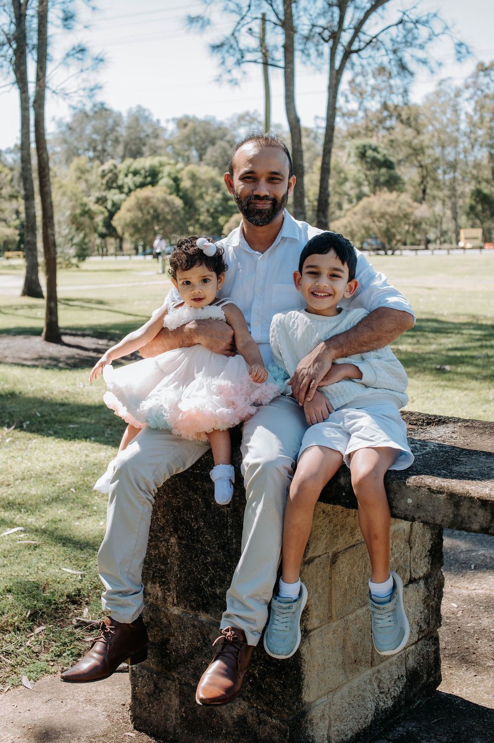 Family members sharing joyful moments together while sitting on a rustic stone wall in a sunny park setting.