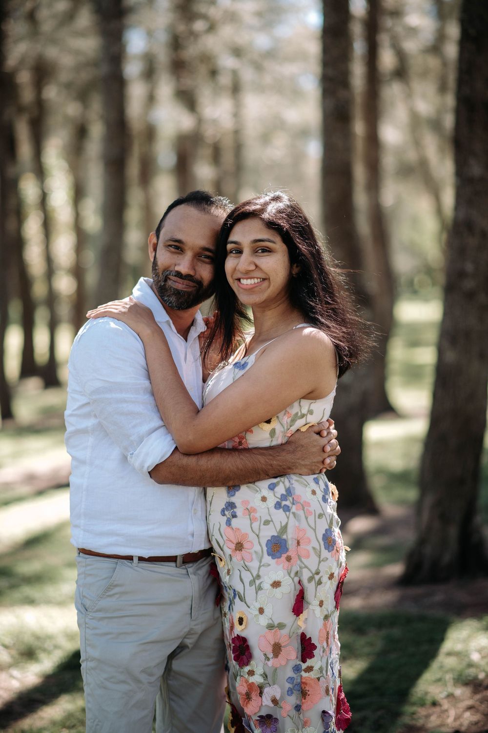 A couple embraces in a sunny forest setting, wearing casual summer attire with floral print dress and white shirt.