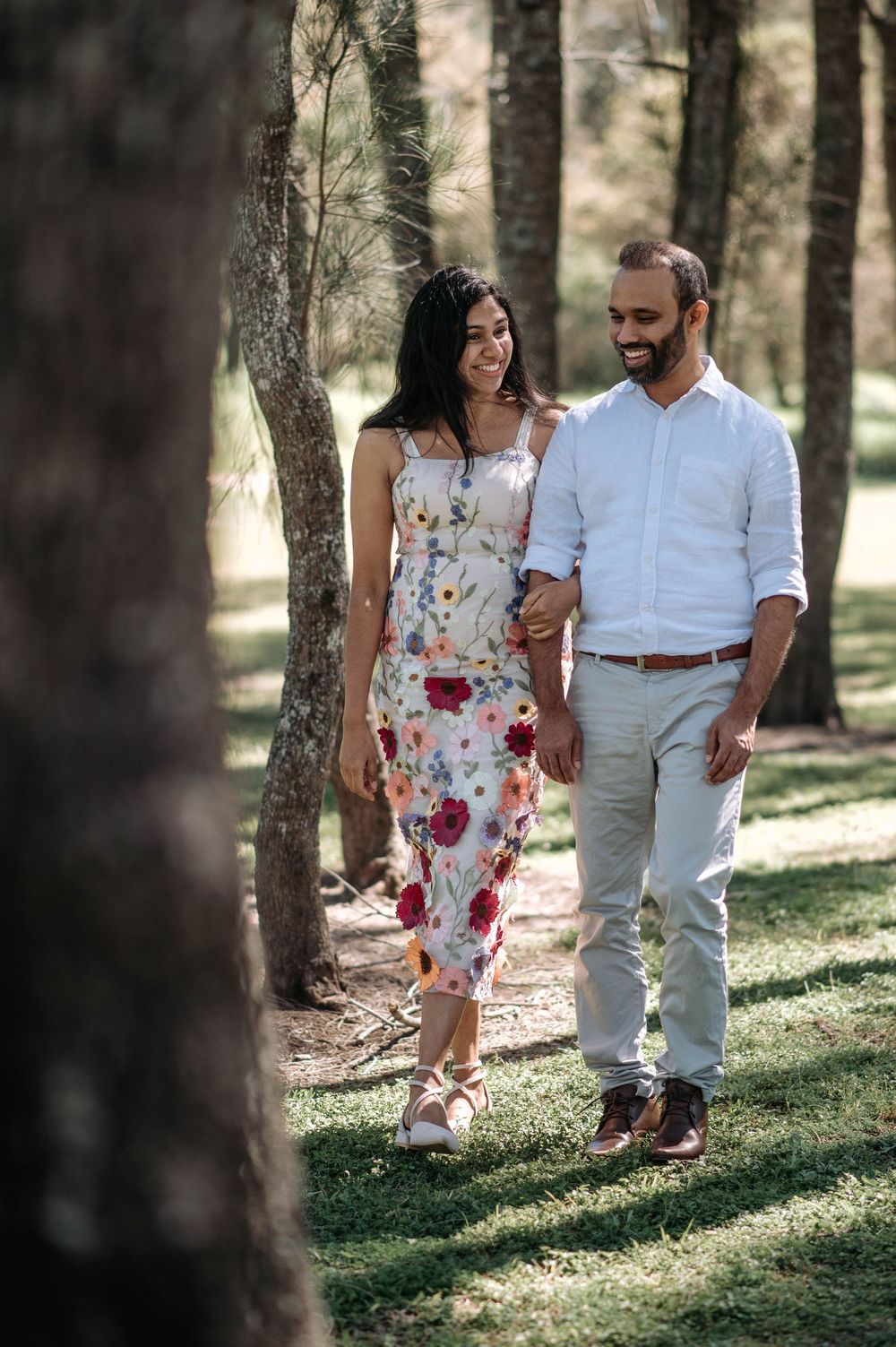 Couple walking together through a sunlit wooded area, with the woman wearing a floral dress.