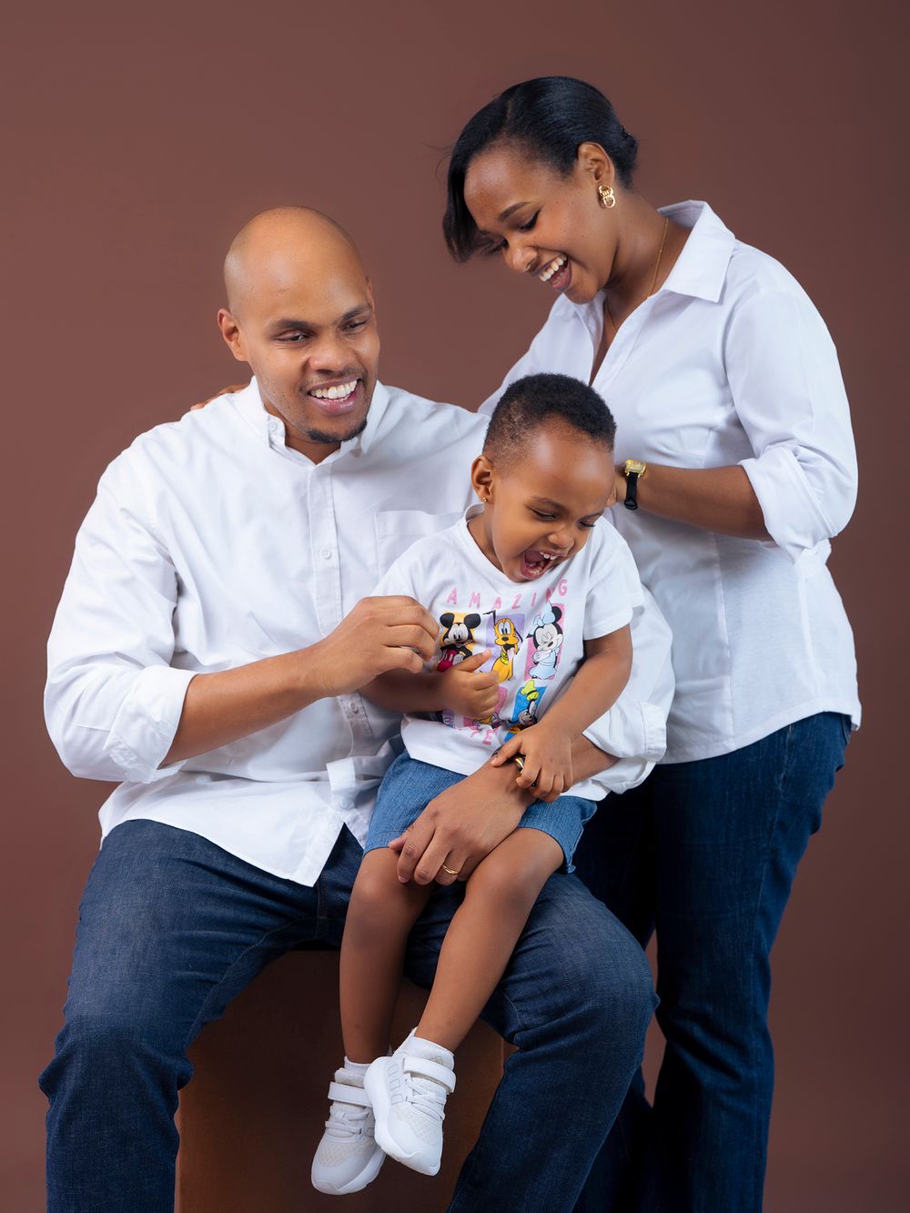 A small family shares a playful moment together against a brown backdrop while wearing white shirts and jeans.