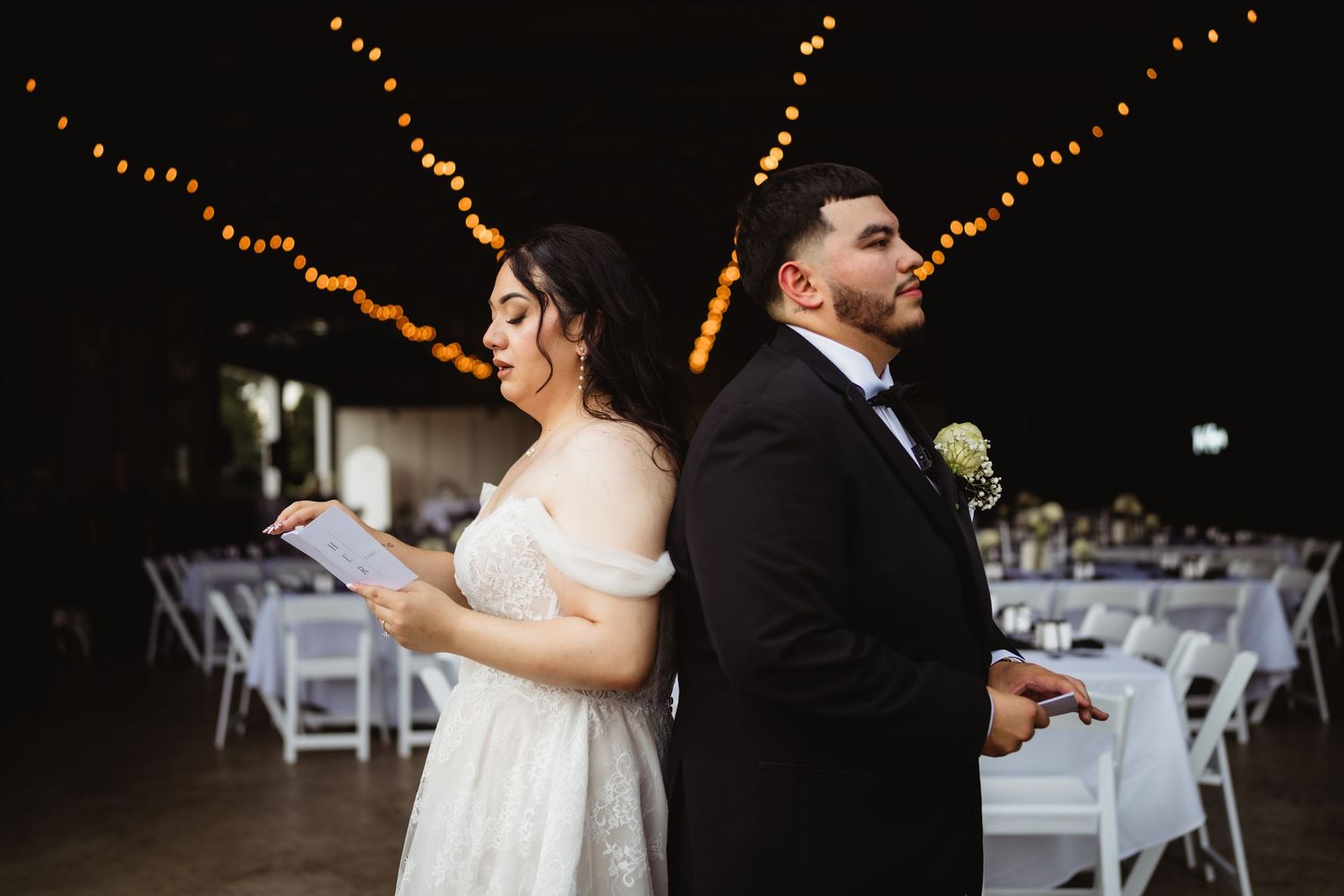 A couple stands back to back in a barn reception venue with string lights and white chairs.