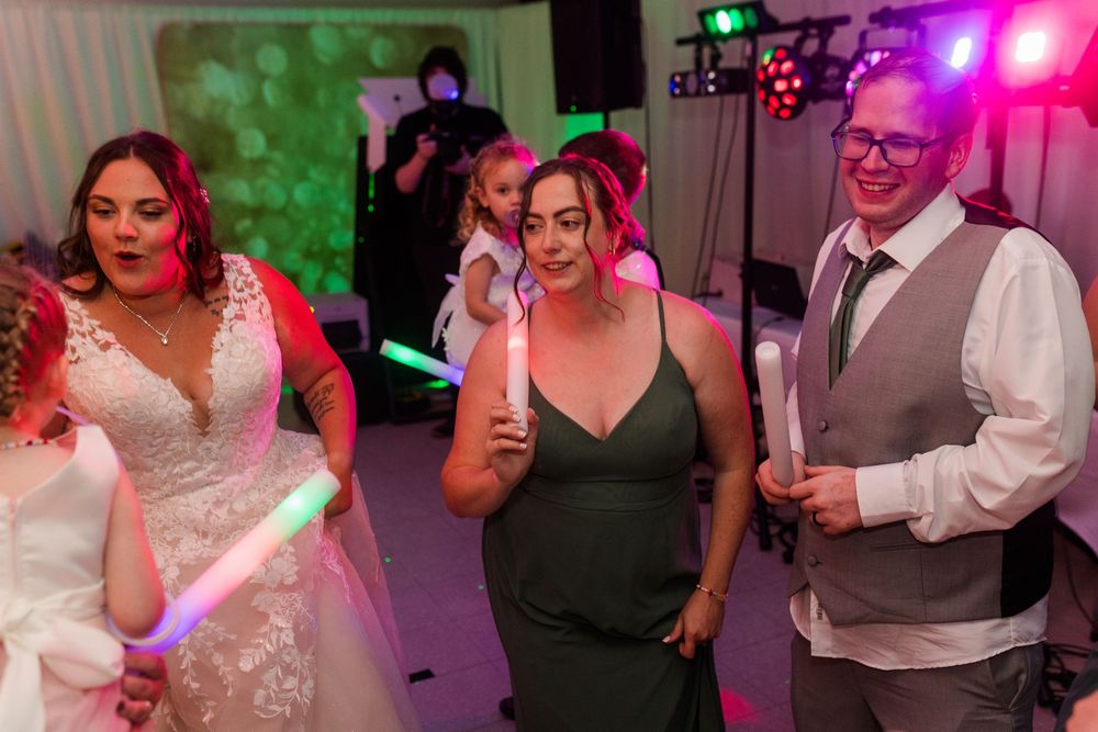 People dancing and celebrating at a wedding reception with colorful purple and green lighting effects.