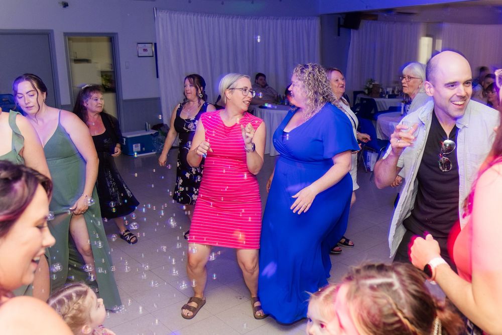 Party guests dance energetically on a dance floor with colorful lighting and bubbles at a celebration event.