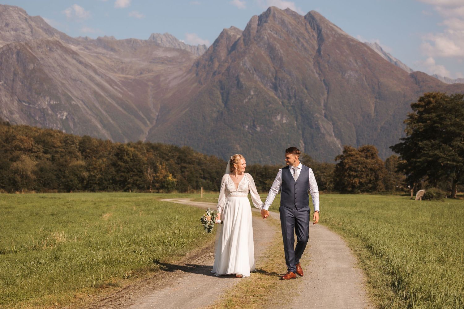 A scenic wedding photoshoot shows silhouettes walking through a dramatic mountain landscape in golden sunlight.