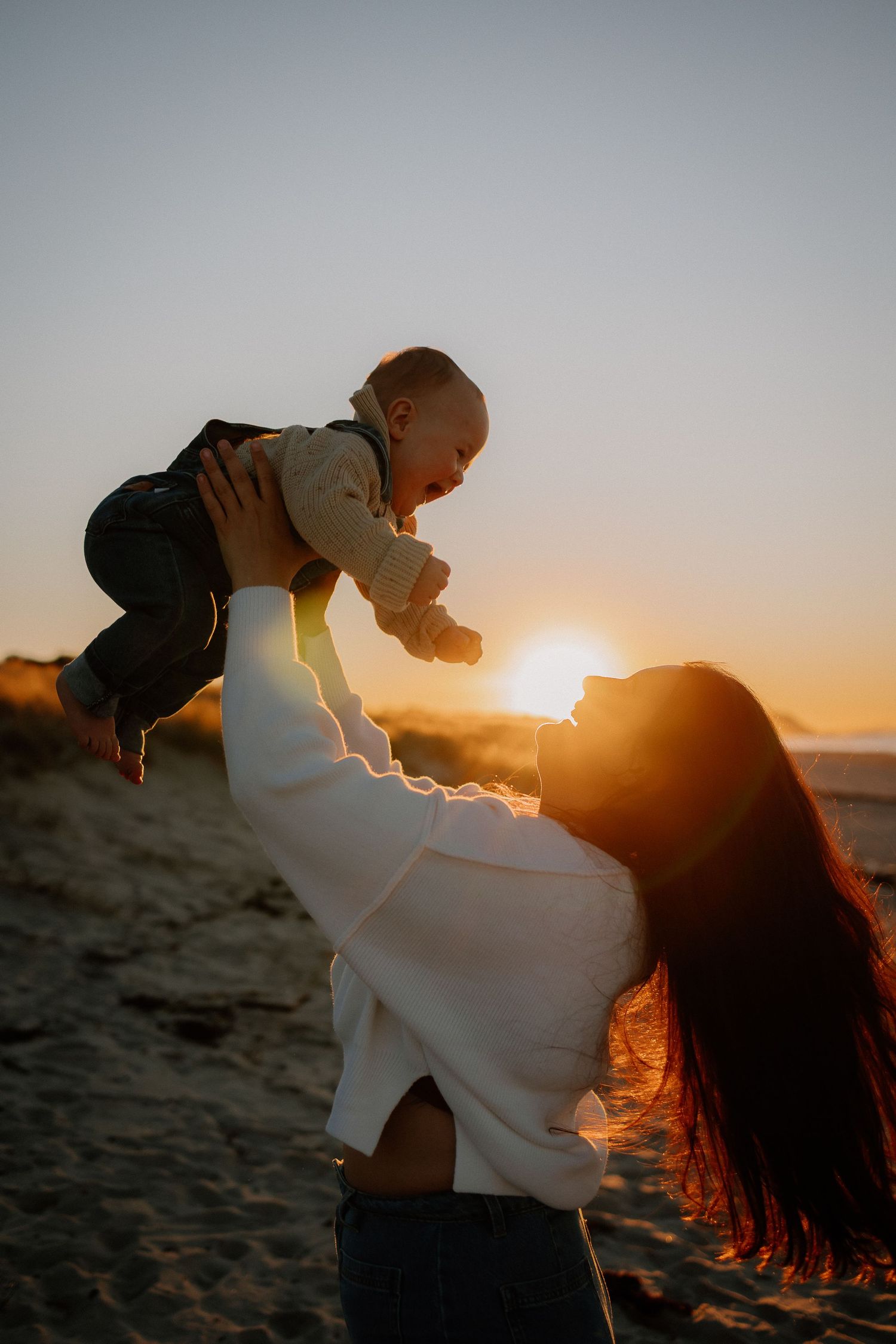 A parent lifts their baby in the air against a golden sunset backdrop on a sandy beach, creating a joyful silhouette.