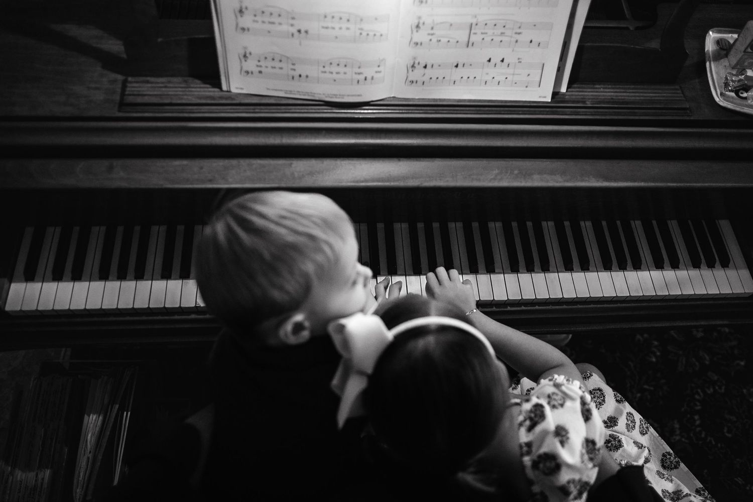 A black and white view of a small child reaching up to play piano keys with sheet music visible above.