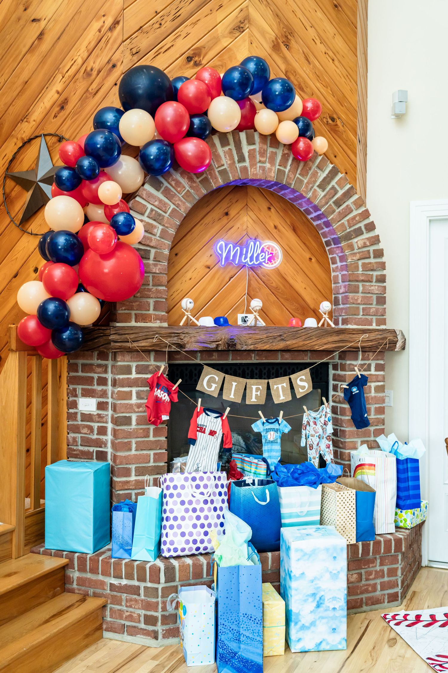 Baby shower display with red and blue balloon arch over brick fireplace with wrapped presents.