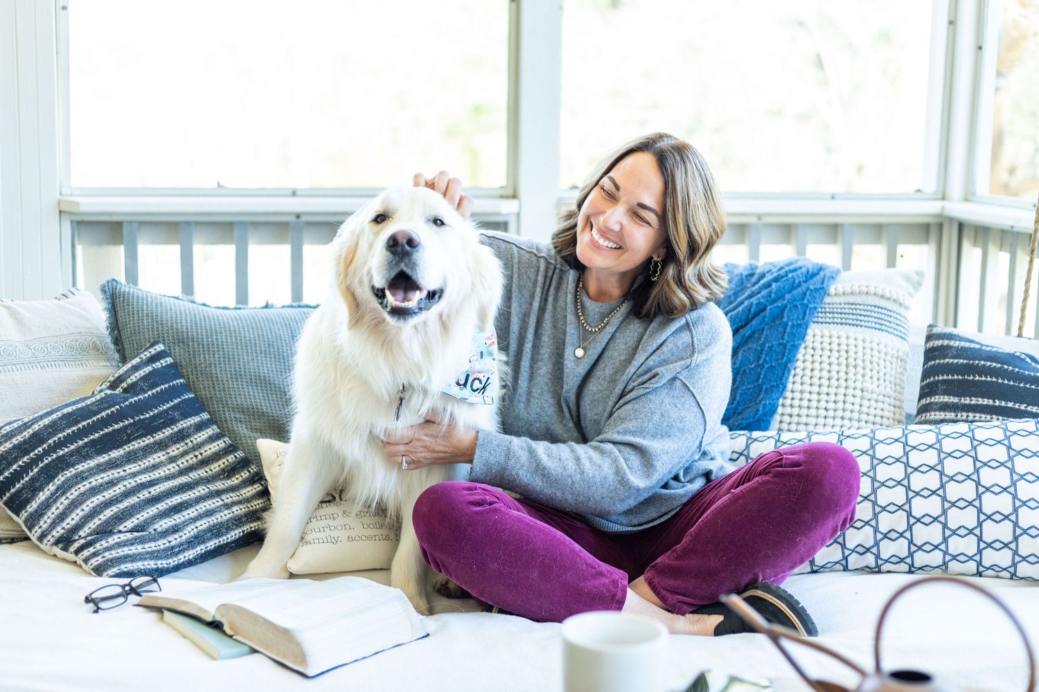 Woman relaxing on a couch with a white dog while working on a laptop in a bright modern living room.