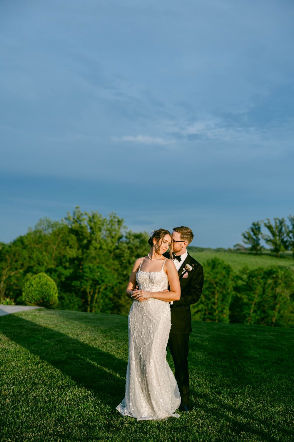 A romantic wedding couple embrace on a lush hilltop during golden hour against a blue sky and green landscape.