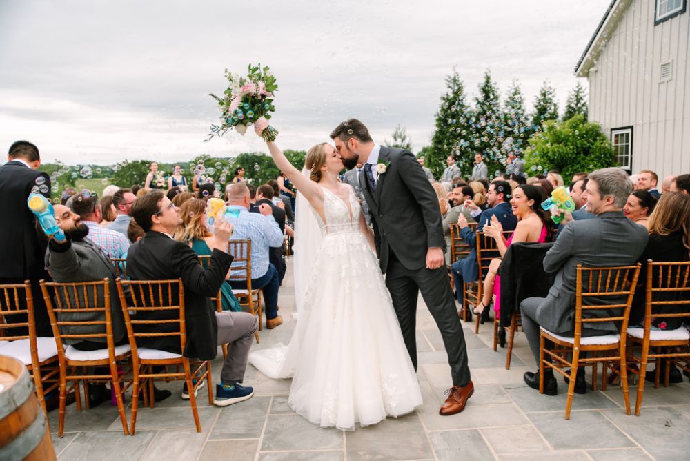 Guests attending an outdoor wedding ceremony with wooden chairs and a rustic barn setting.