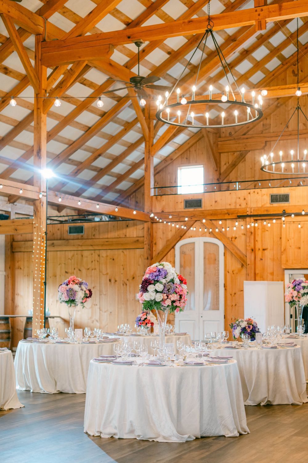 Elegant barn reception with wooden beams, chandeliers, and white table settings with floral centerpieces.