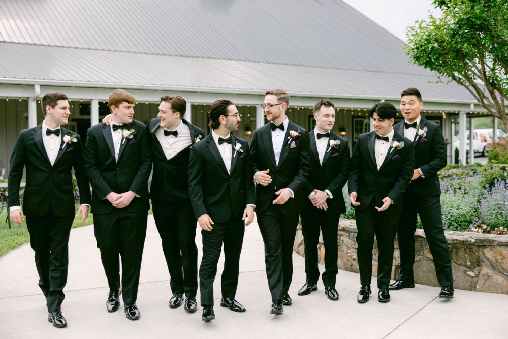 Groomsmen in black tuxedos walking in line formation outdoors.