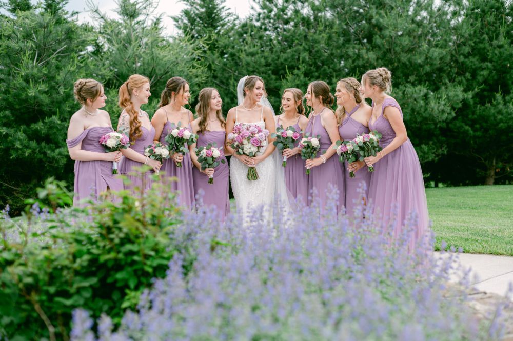 Bridal party in lavender dresses poses together in garden with purple flowers in foreground.