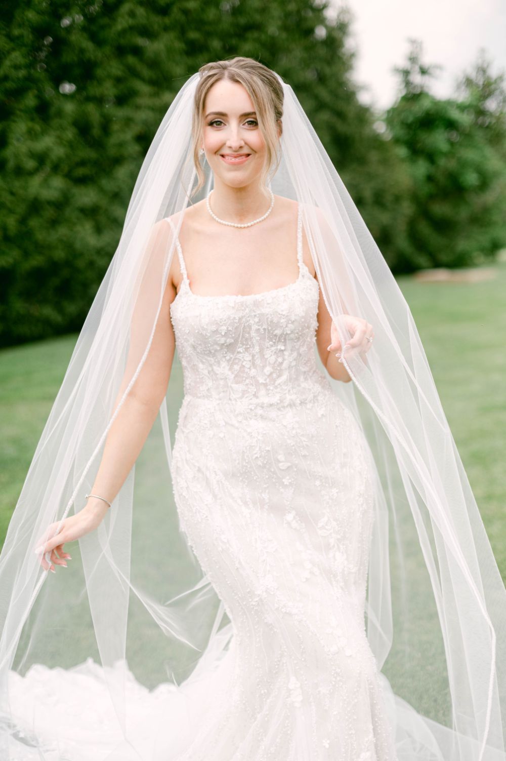 Elegant white lace wedding dress with delicate straps and flowing veil in an outdoor garden setting.