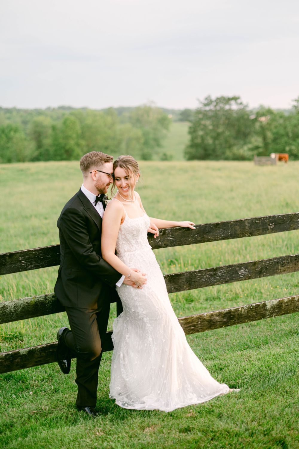 A couple in wedding attire poses together by a rustic wooden fence in a green field.