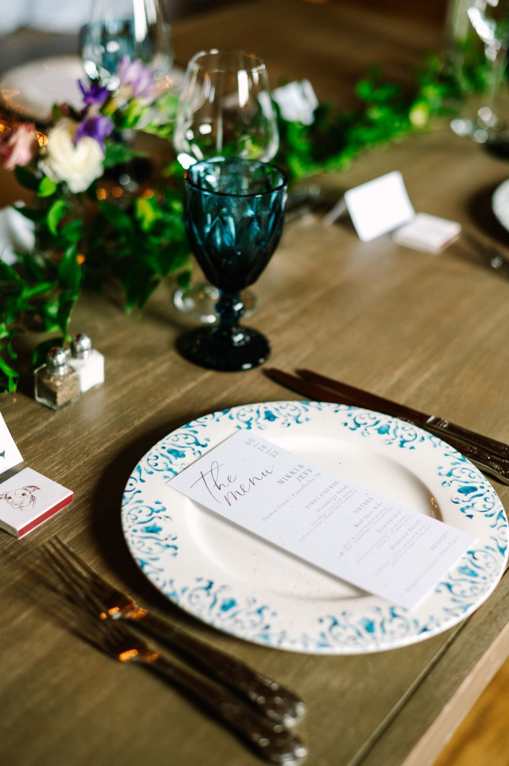 Decorative wedding place settings with blue-trimmed plates and elegant greenery on a rustic wooden table.