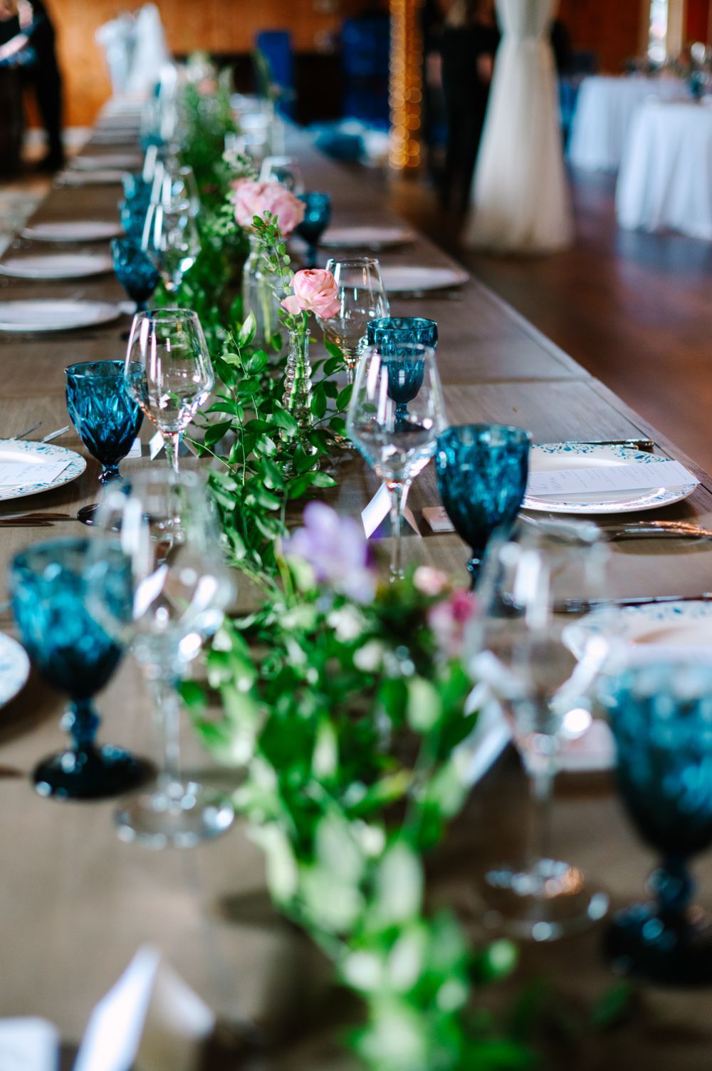 Wedding reception table decorated with blue glassware, greenery garlands, and pink roses creates an elegant tablescape.