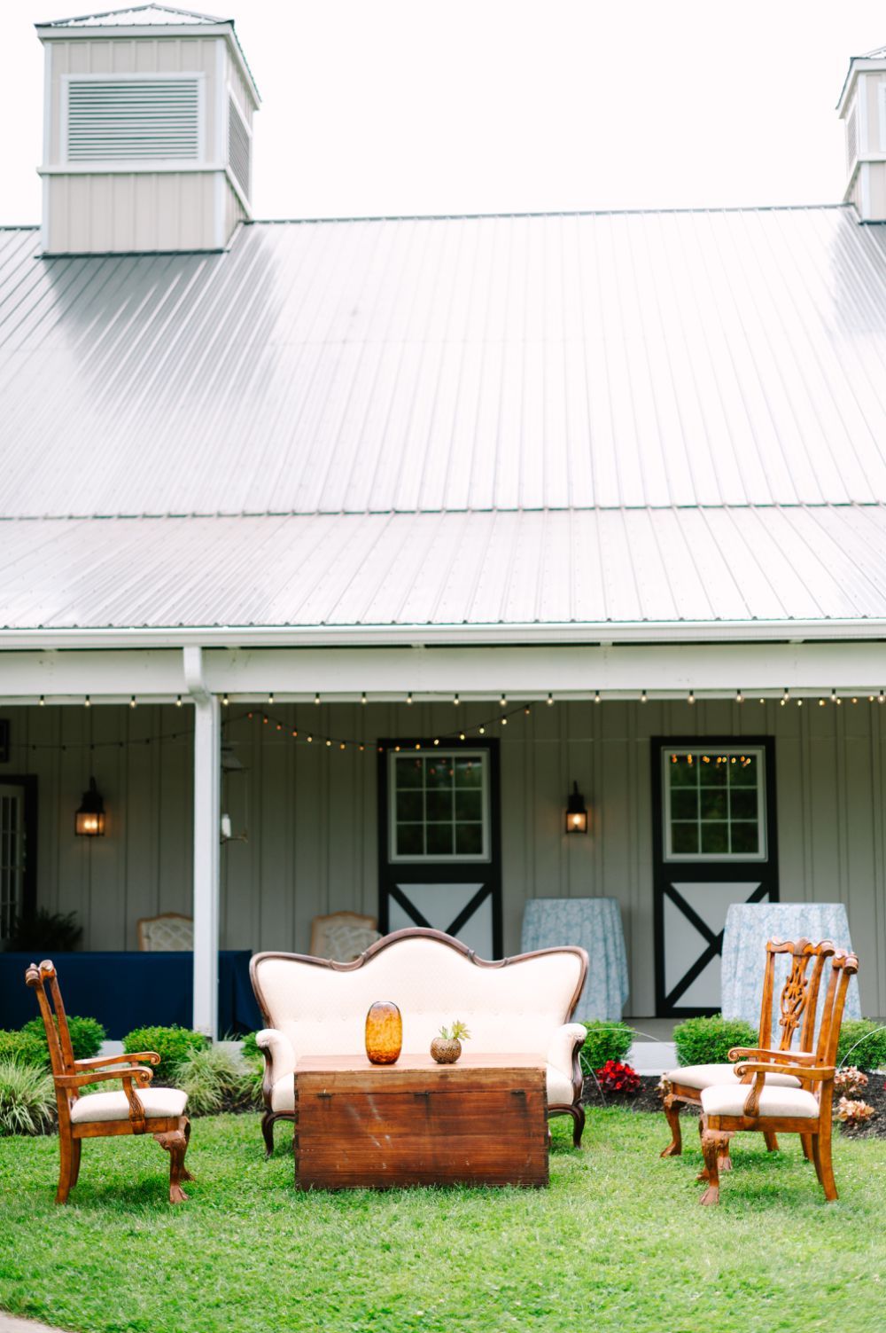 Vintage wooden furniture including chairs and tables arranged on a lawn in front of a white farmhouse.