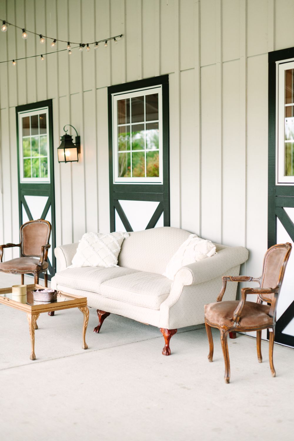 Elegant veranda with white cushioned seating, wooden chairs and string lights creating a cozy outdoor living space.