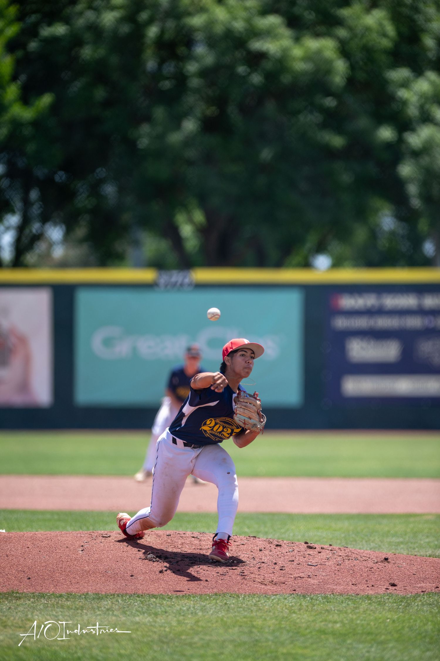 Baseball pitcher in red cap follows through on delivery motion during outdoor game on sunny afternoon.