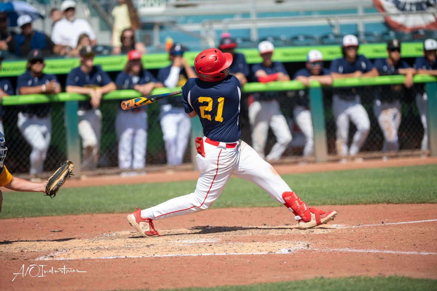 Baseball batter in dark uniform with red accents swings at home plate during daytime game.