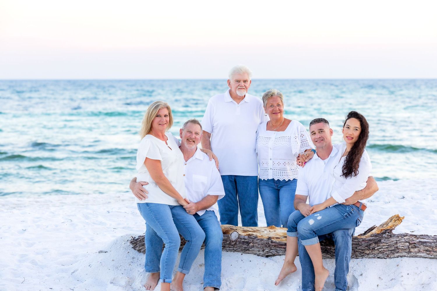 Family group dressed in white and blue sits together on rocks at the beach during sunset.