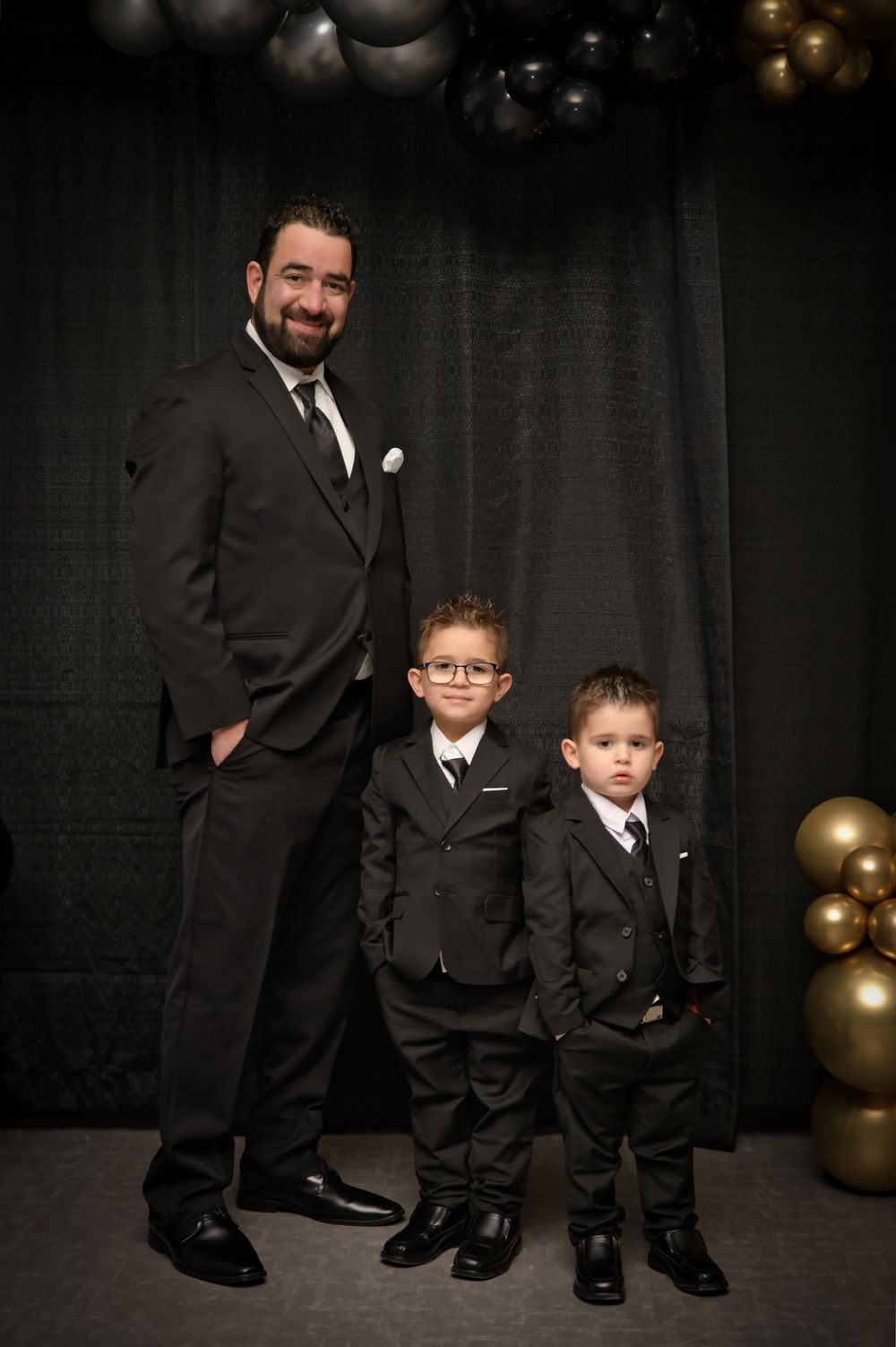Father and two young boys in matching black suits pose together against dark backdrop with gold balloons.