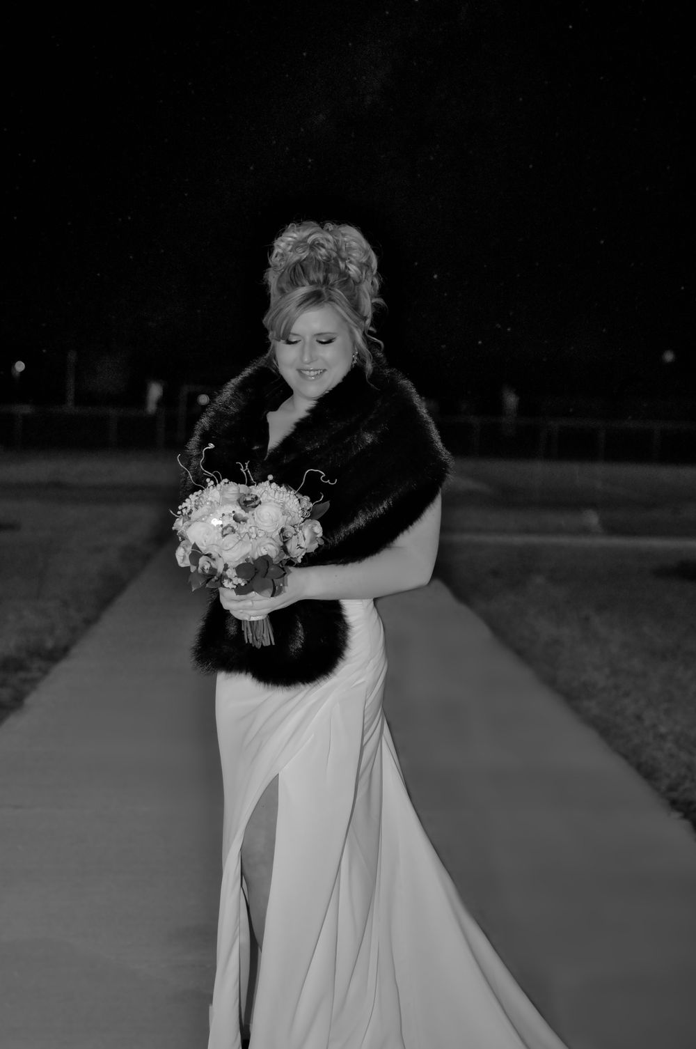 A bride in a white dress with a side slit wears a black fur wrap while holding a bouquet outdoors at night.