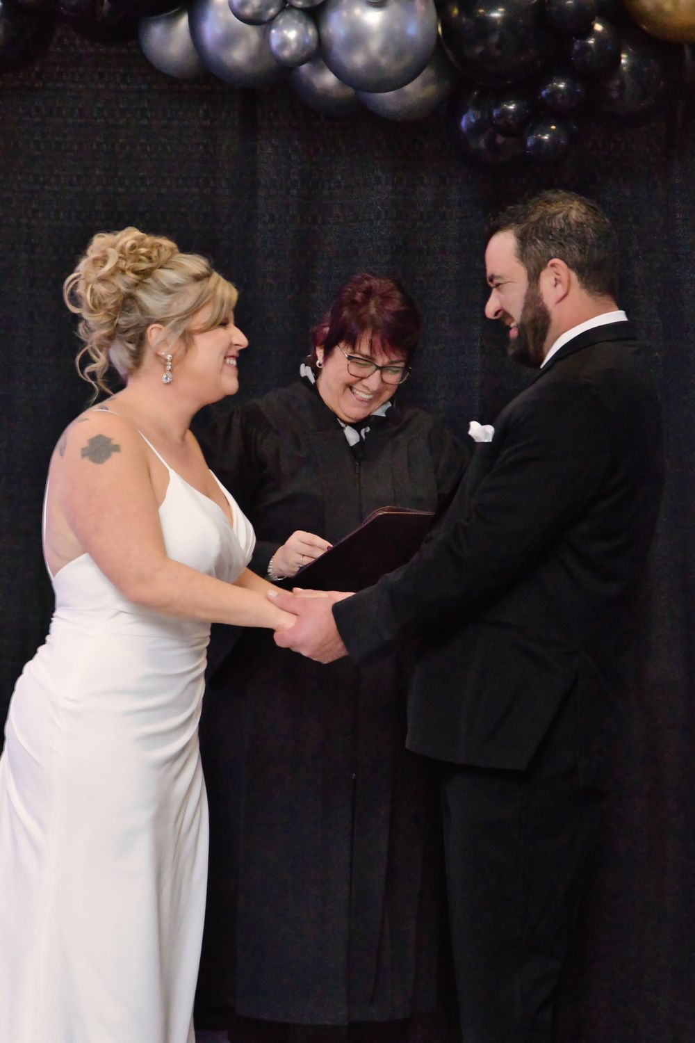 A wedding ceremony takes place indoors with the couple exchanging vows under purple balloon decorations.