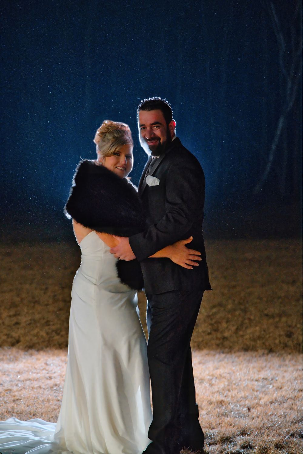 Couple dancing at night in a field, bride in white dress and groom in black suit sharing an intimate embrace.