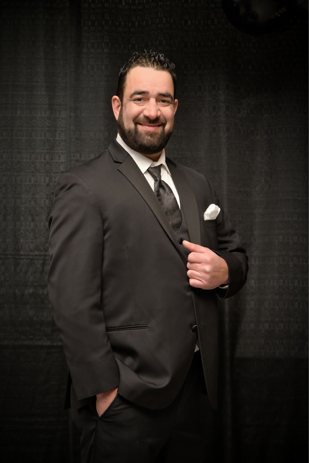 Professional studio portrait of bearded person in dark suit with hand in pocket against dramatic black background.