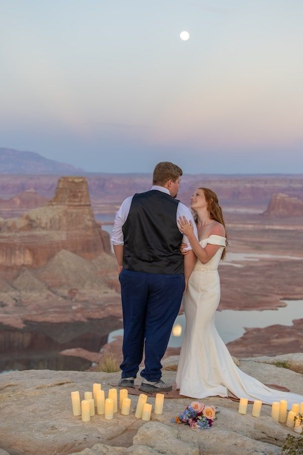 Couple in formal wear embraces at scenic desert overlook surrounded by glowing candles at twilight with full moon rising.