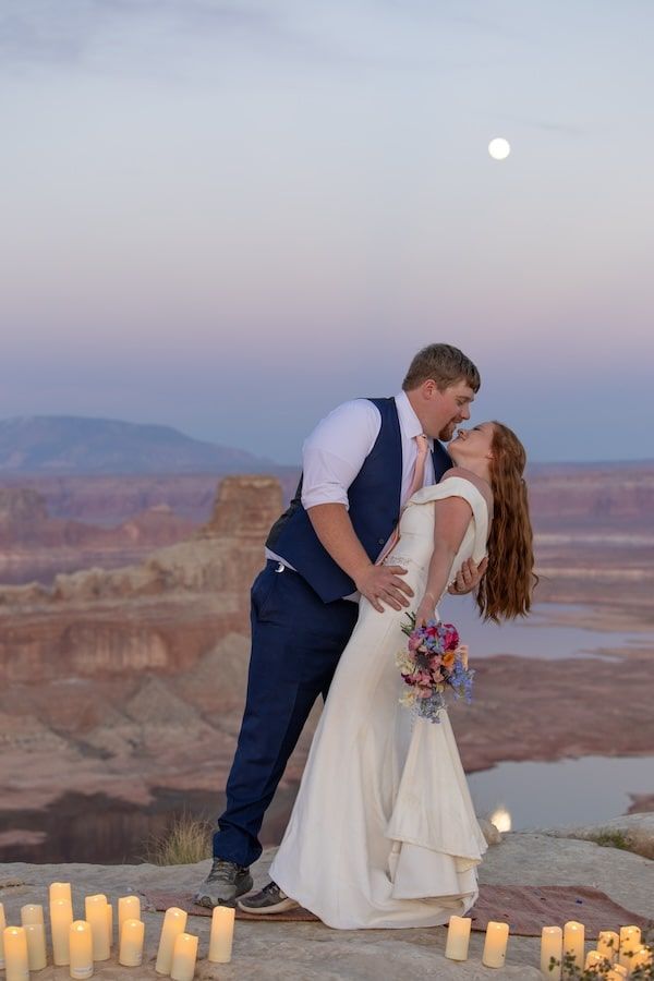 A couple shares a romantic kiss against a stunning desert canyon backdrop with glowing candles at sunset.
