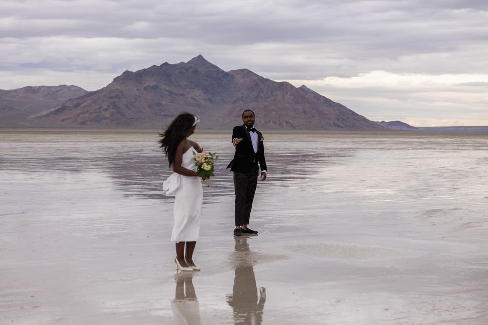 Two people stand on the reflective surface of Bonneville Salt Flats in Utah during a dramatic overcast sunset.