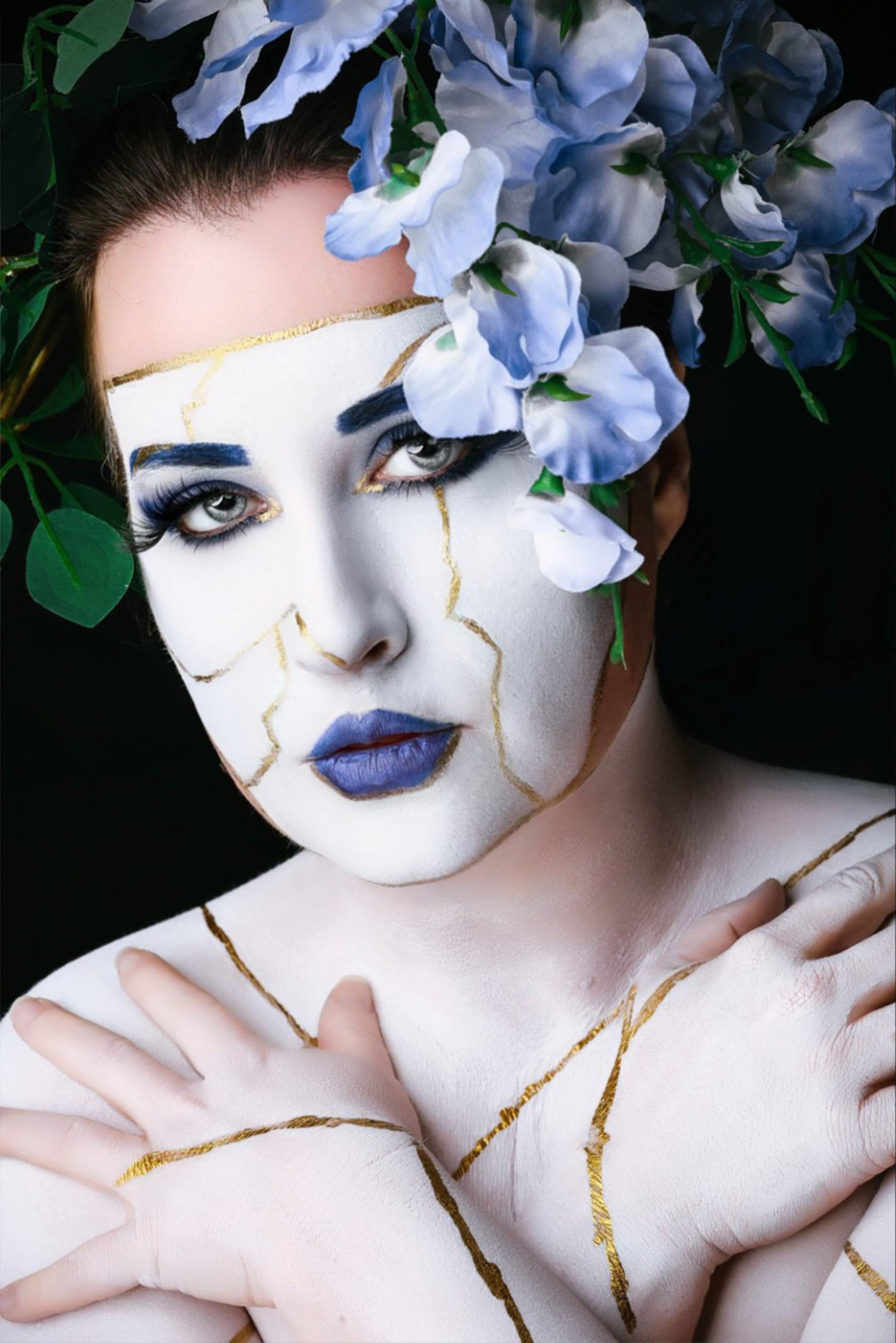 Woman with white face paint, gold kintsugi cracks, blue lips, and floral crown of blue flowers against dark background.