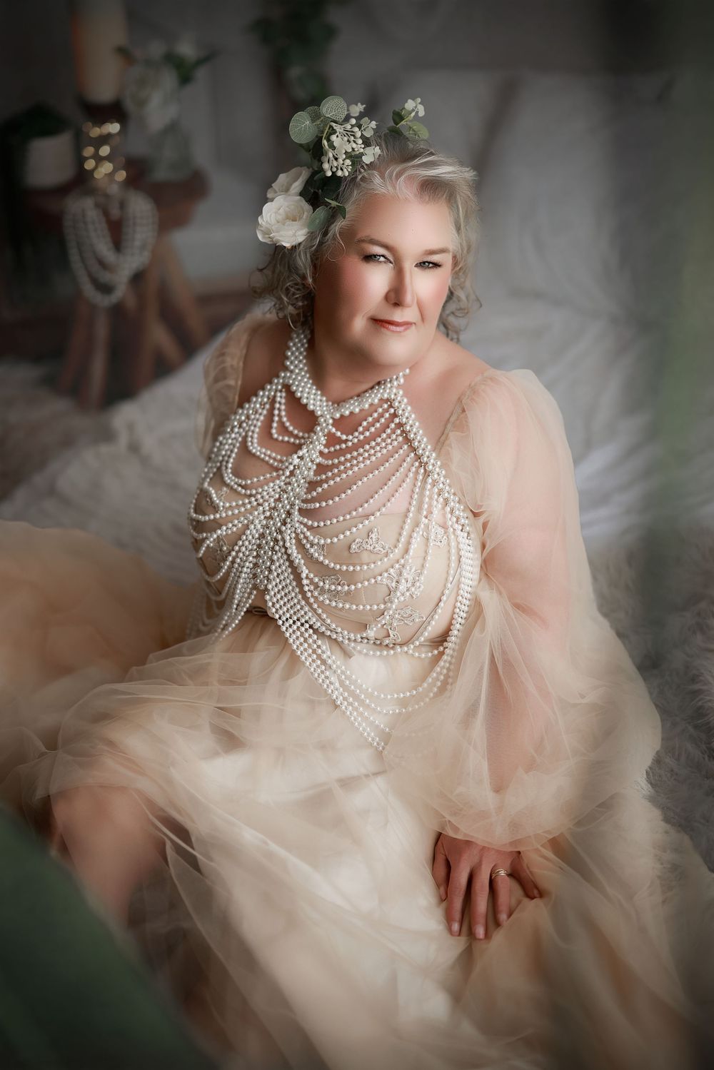 Elegant older woman in blush tulle gown adorned with pearl draping necklace and floral hair accessories poses gracefully.