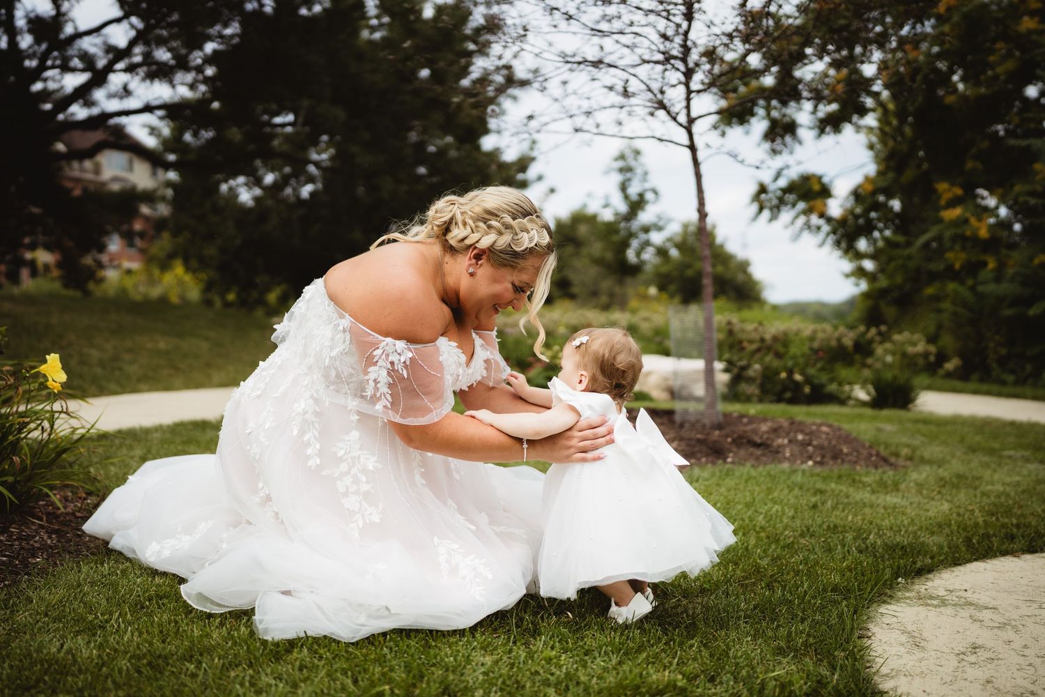 Small spotted dog in a bowtie lovingly rests near a flowing white wedding dress on green grass.