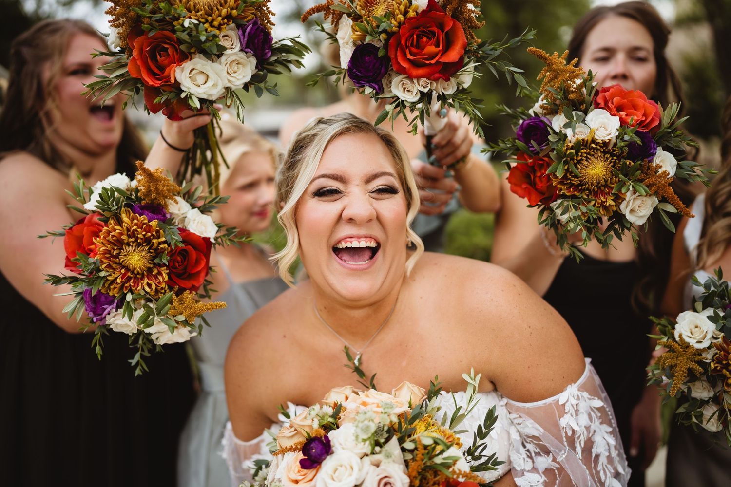 Joyful wedding party holding red and white floral bouquets while laughing and celebrating outdoors.