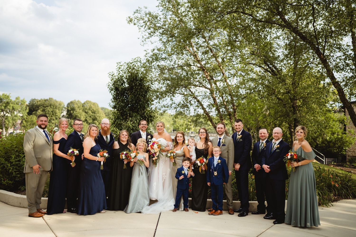 A wedding party group poses outdoors on a sunny day with trees and greenery in the background.