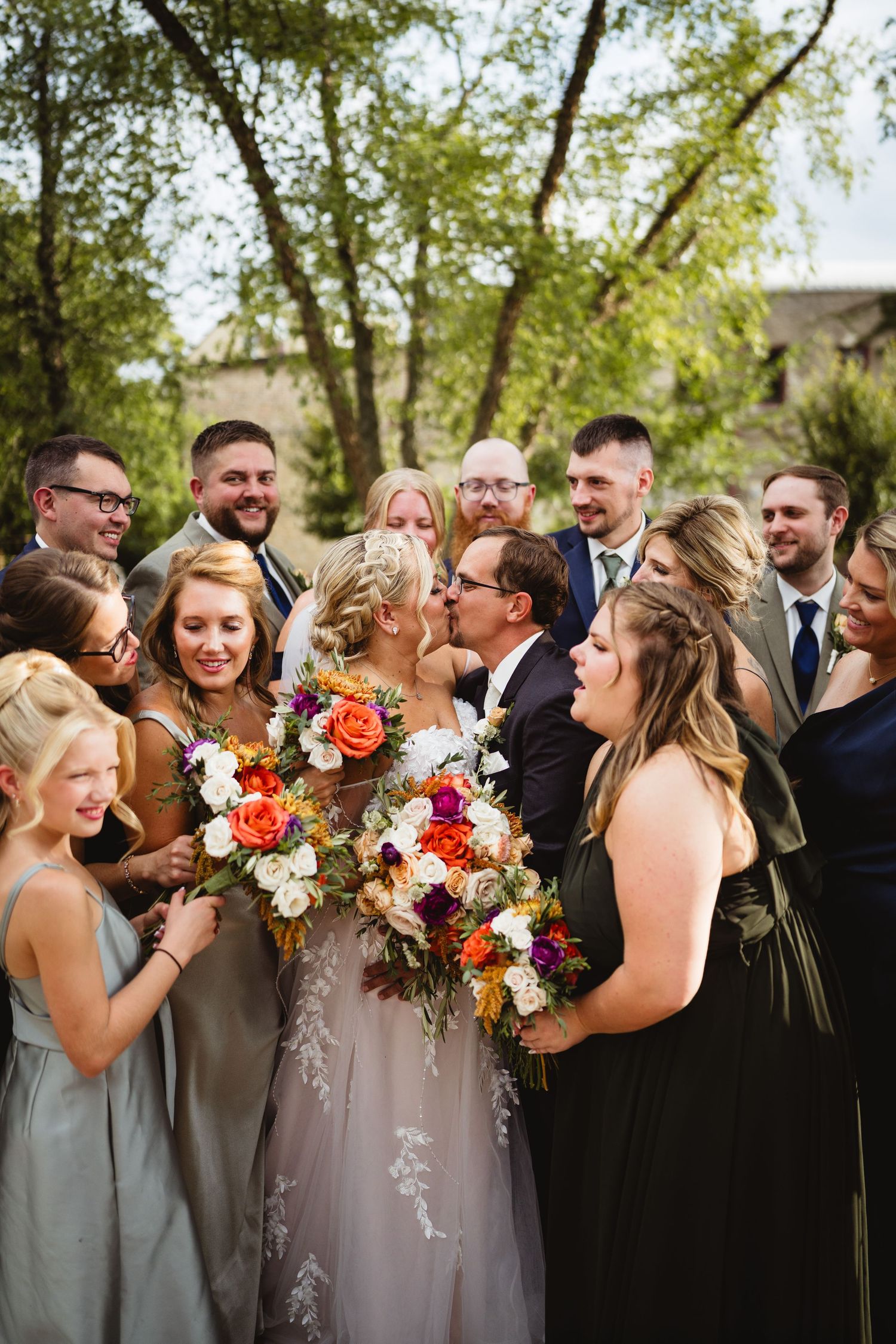 A joyful wedding party laughs together while holding colorful fall bouquets with orange and white flowers outdoors.
