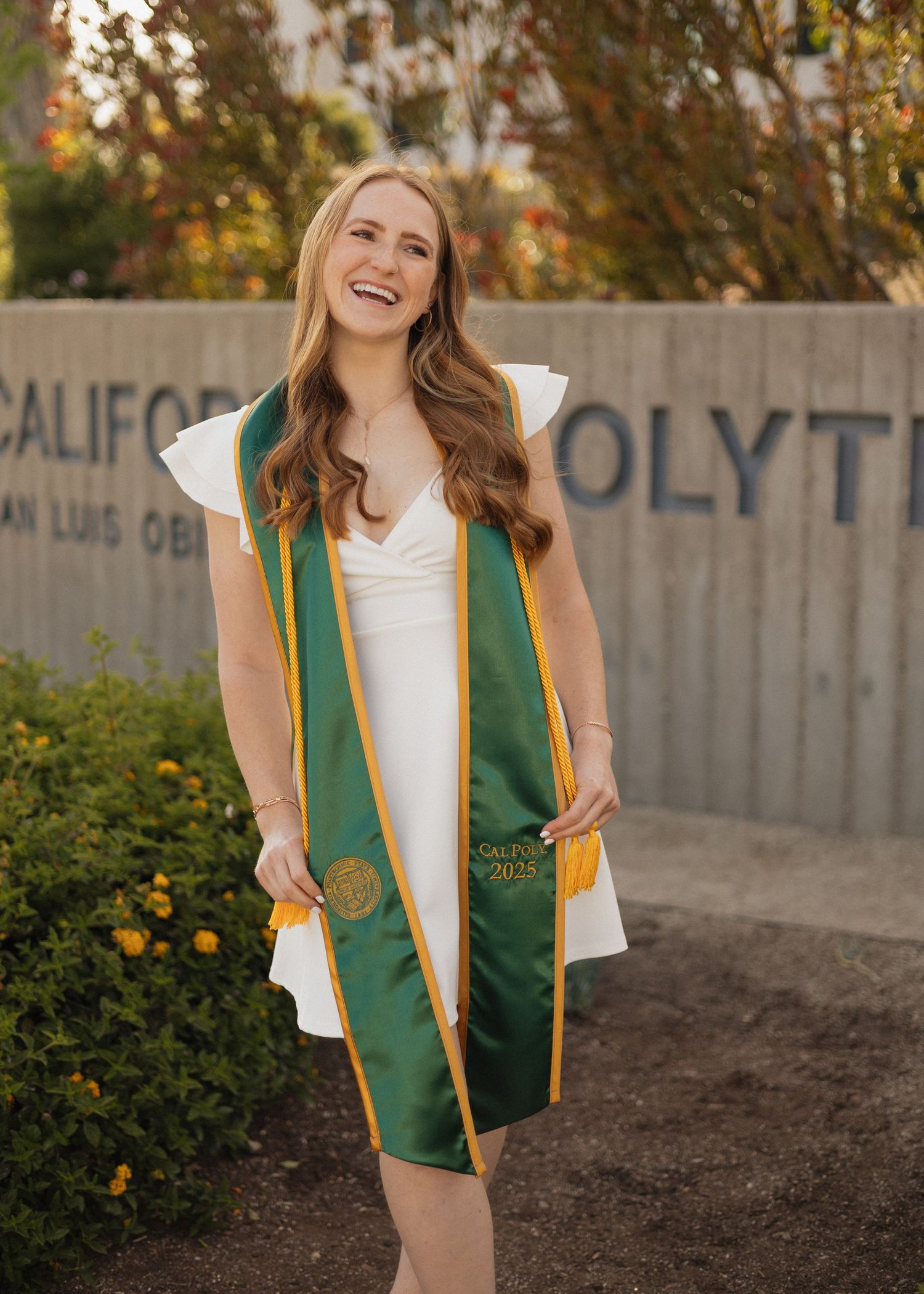 Cal Poly SLO graduate in white dress and green stole smiles in front of college signage.