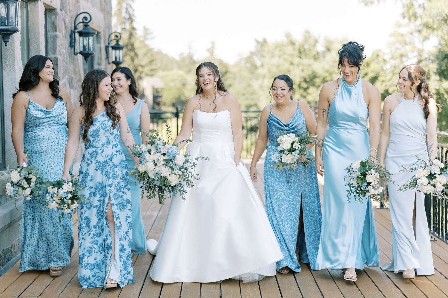 Wedding party in light blue bridesmaid dresses and white wedding gown pose on stone porch during outdoor celebration.