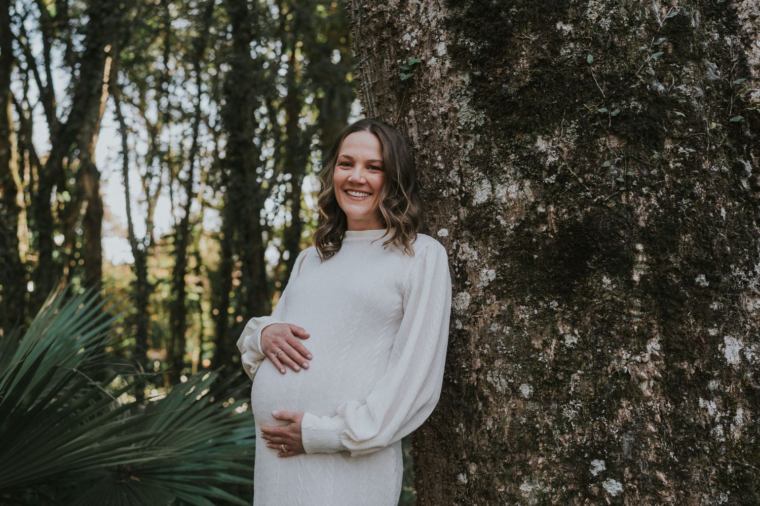 Outdoor maternity portrait against a tree trunk in a natural woodland setting.