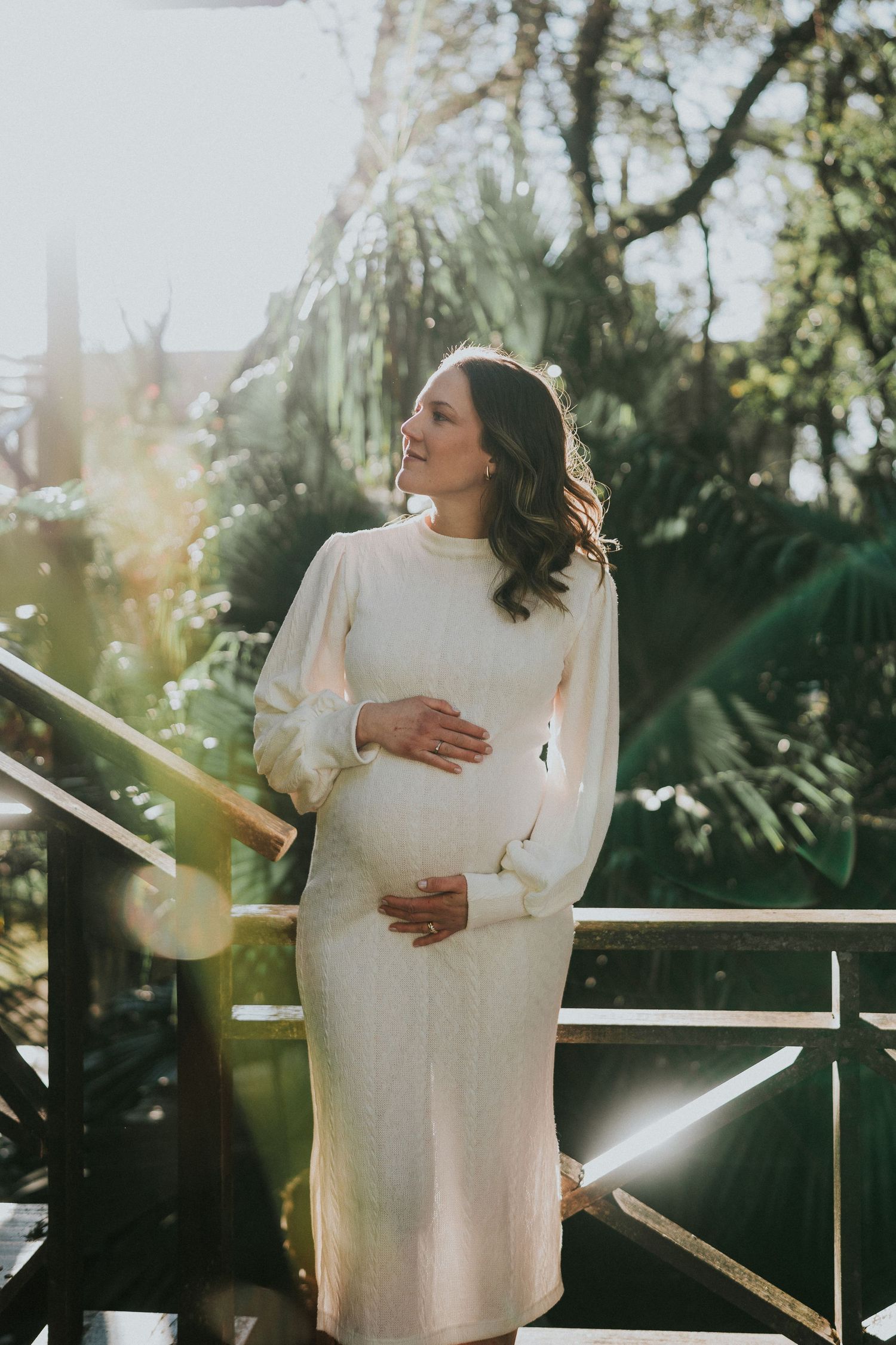 Dreamy maternity portrait on a balcony with sunlit tropical foliage in the background.